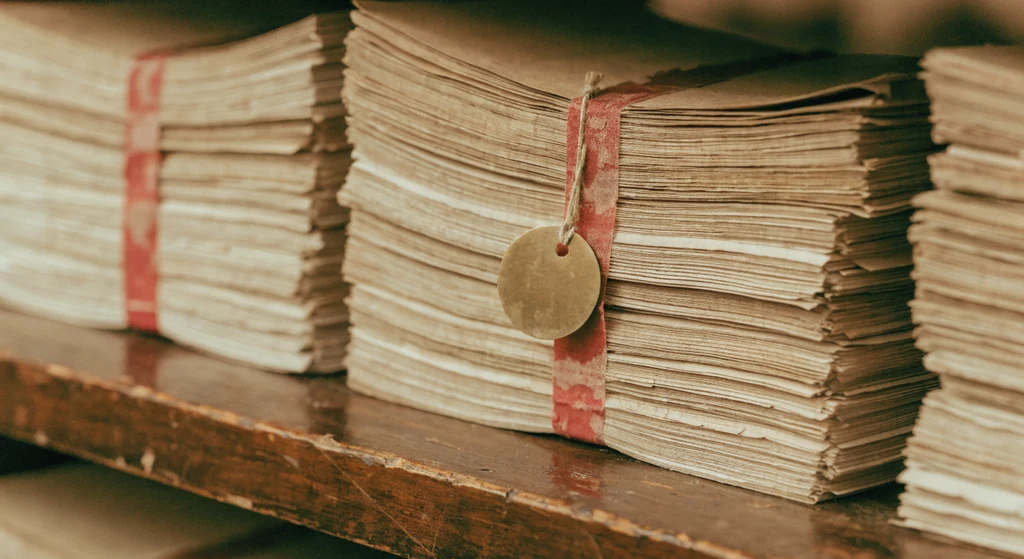Aged document bundles tied with faded red cotton tape stacked on a dark wood shelf, a plain brass tag hanging from one
