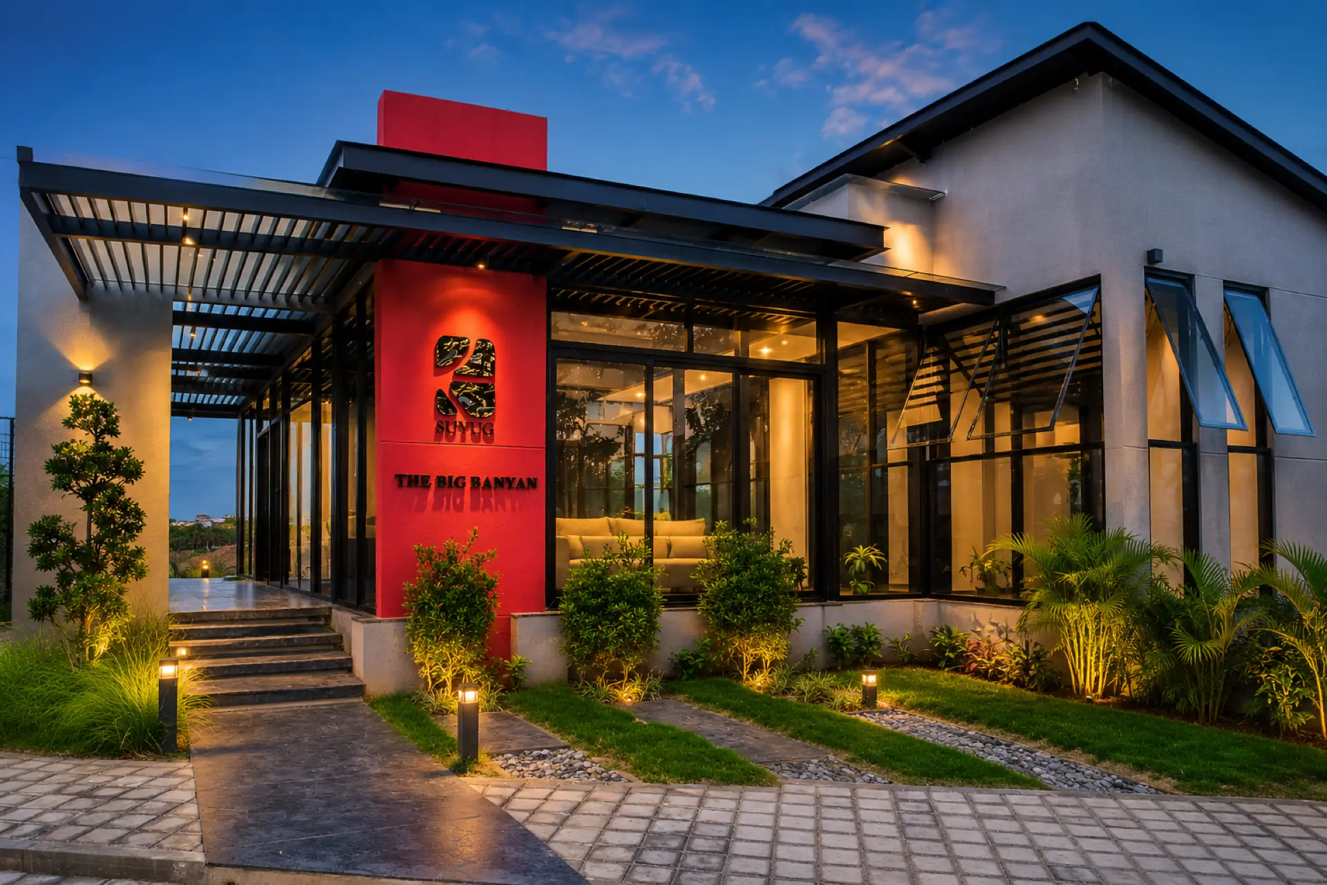 The Big Banyan marketing office at dusk: a red column carrying the SUYUG mark and The Big Banyan name, glass walls glowing warm under a pergola.